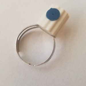 May include: A ring featuring a white, rectangular top with a blue circular accent. The ring band is silver-colored and has a textured design. The ring is set against a plain white background.