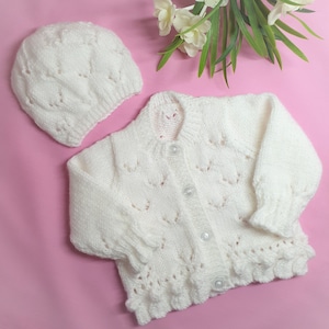 Könnte beinhalten: Weißes gestricktes Baby-Cardigan und passende Mütze mit zartem Spitzenmuster. Das Cardigan hat einen Knopfverschluss und die Mütze ist ein einfacher Beanie-Stil.
