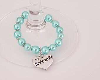 May include: A light blue glass charm with a silver heart charm that says "Bride to Be". The charm is made of pearls and is designed to be placed around a wine glass.