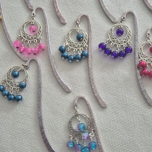 Può includere: Un set di nove segnalibri in metallo argentato con ciondoli pendenti. Ogni ciondolo presenta un design circolare con una frangia di perline. Le perline sono di vari colori, tra cui blu, viola, rosa e turchese.