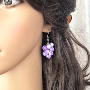 Puede incluir: Pendientes de cuentas de vidrio morado y blanco con un gancho plateado. Los pendientes son colgantes y tienen un racimo de cuentas.