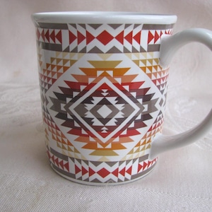 May include: White ceramic mug with a geometric pattern in shades of red, orange, brown, and gray. The design features diamond and triangle shapes. The mug has a white handle and rim.