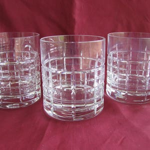 May include: Three clear, crystal lowball glasses. Each glass features a grid-like pattern around the lower half. The glasses are cylindrical with a thick base and are displayed on a maroon surface.