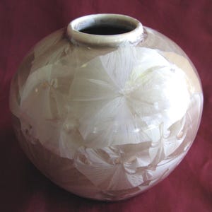 May include: A round ceramic vase with a smooth, glossy finish. The surface features a crystalline glaze pattern with large, white, star-shaped crystal formations set against a soft, muted mauve and light brown background color palette.