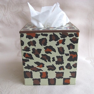 May include: A decorative tissue box with a leopard print design. The box is square with a light green background and brown and orange spots. The top and trim are brown. White tissues are visible.