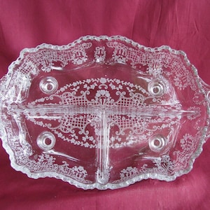 May include: A clear glass divided serving tray featuring an etched white floral and lattice pattern. The oval-shaped dish has two side handles, a scalloped rim, and four small circular feet on the base, creating three distinct sections for food.