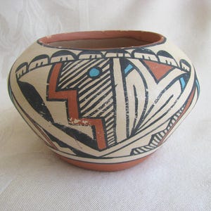 May include: A round, hand-painted ceramic pot with a cream-colored base. The pot features geometric designs in black, red, and blue, including a zigzag pattern and stylized shapes. The rim and base are a reddish-brown color.