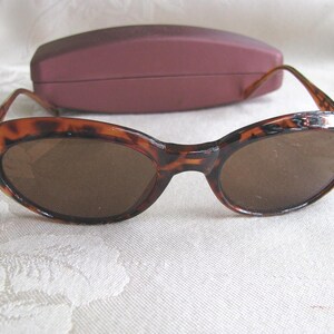 May include: Brown tortoiseshell sunglasses with oval lenses and a matching maroon case. The sunglasses have a classic design with a slightly curved frame and dark lenses. The case is rectangular and provides protection for the eyewear.