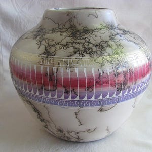 May include: A handcrafted ceramic vase with a wide body and a narrow neck. The vase features a white base with black, swirling patterns. A band of pink and purple vertical lines is accented by a silver geometric design. The top is a light gray color.