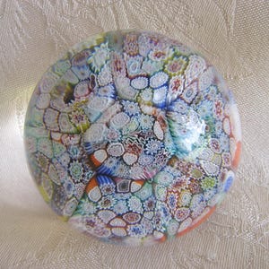 May include: A spherical glass paperweight featuring a dense millefiori pattern with numerous small, intricate floral and geometric shapes in shades of blue, white, green, orange, and yellow embedded within the clear, polished glass surface.