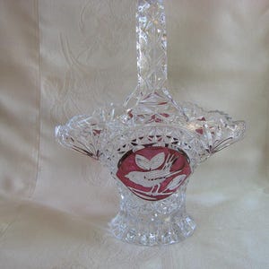 May include: A clear crystal basket with a handle, featuring a cut glass design. The basket has a red interior with a white bird and leaf design. The basket is likely used for decorative purposes.