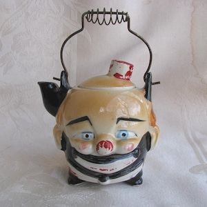 May include: A vintage ceramic teapot shaped like a clown's head. The teapot features a tan face with blue eyes, a red nose, and a black and white striped outfit. It has a metal handle and a black spout.