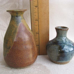 May include: Two small ceramic vases. The larger vase is approximately 4 inches tall, with a brown and green color scheme. The smaller vase is about 2 inches tall, with a blue and gray glaze. A wooden ruler is placed between the vases.