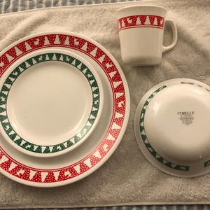 May include: A white dinnerware set consisting of a large plate, a smaller plate, a bowl, and a mug. The large plate and mug feature a red border with white trees and deer. The smaller plate and bowl feature a green border with white trees and deer.