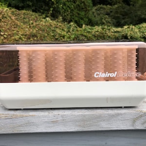 May include: A Clairol Deluxe electric hair setter featuring a white base and a translucent brown lid. Inside the unit, a series of light peach-colored hair rollers with textured surfaces are arranged in rows, accompanied by a white power cord.