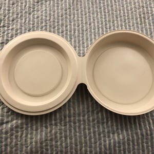 May include: A white, double-bowl pet feeder made of plastic. The feeder has two circular bowls connected by a bridge. Each bowl has a raised rim and a circular indentation in the center. The feeder is designed for pets.