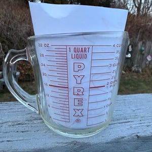 May include: A clear Pyrex measuring cup with a handle, featuring red measurement markings for ounces, cups, and quarts. The text "PYREX" is prominently displayed. The cup is filled with a white sheet of paper.