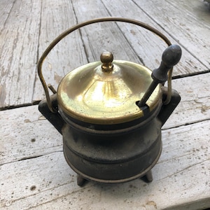 May include: A small, round, black cast iron pot with three short legs and a brass lid featuring a central knob. The pot includes a curved brass handle and a dark wooden spoon resting in a small notch on the side of the lid, creating a functional set.