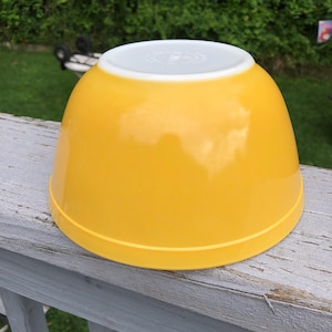 May include: A bright yellow mixing bowl with a smooth, glossy finish and a white interior rim. The bowl is inverted, showing a circular base with embossed text markings visible on the white center portion of the bottom surface of the bowl.
