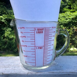 May include: A clear glass measuring cup with a handle and a pouring spout. The exterior features red printed markings indicating measurements in cups, ounces, 1 quart liquid, and 1 pint. A white paper insert is placed inside the glass container.