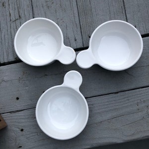 May include: Three white ceramic bowls with a handle-like extension. The bowls are round with a smooth, glossy finish. The bowls are arranged on a weathered gray wooden surface.