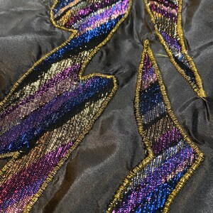 May include: Close-up of a black fabric with a gold and iridescent purple and blue beaded design. The design is in the shape of a lightning bolt.
