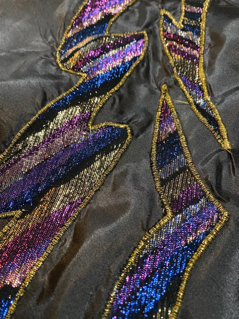 May include: Close-up of a black fabric with a gold and iridescent purple and blue beaded design. The design is in the shape of a lightning bolt.