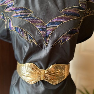 May include: A black silk dress with a gold mesh belt featuring a bow. The dress has a unique embroidered design on the neckline with gold and purple thread.