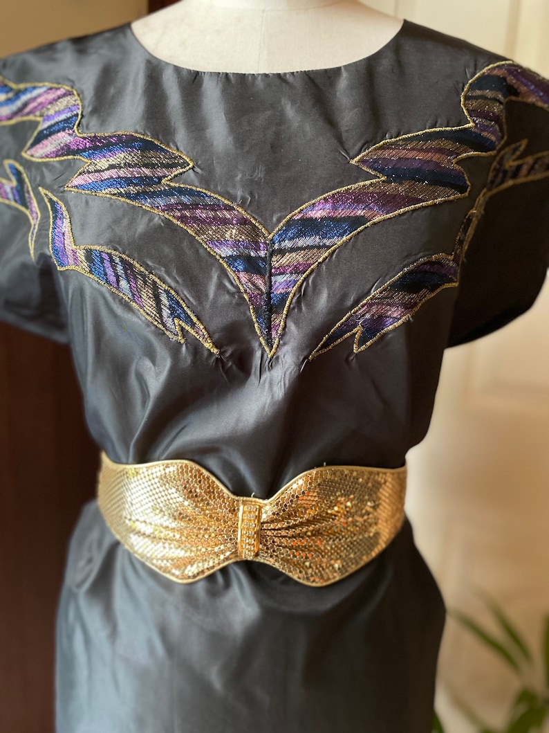 May include: A black silk dress with a gold mesh belt featuring a bow. The dress has a unique embroidered design on the neckline with gold and purple thread.