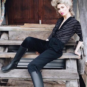 May include: A person wearing a black and white striped top, black trousers, and black boots, sitting on wooden steps. The top has a V-neck and three-quarter sleeves. The boots are knee-high.