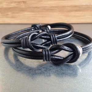 Könnte beinhalten: Zwei schwarze Lederarmbänder mit silbernen Akzenten. Jedes Armband hat ein geknotetes Design.