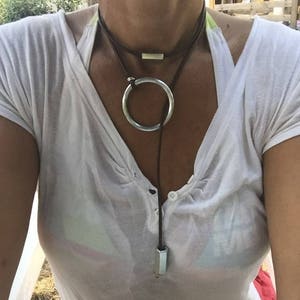 Puede incluir: Un collar con un colgante circular de plata en un cordón de cuero marrón. El collar tiene una pequeña barra de plata en la parte superior y un amuleto hexagonal de plata en la parte inferior.