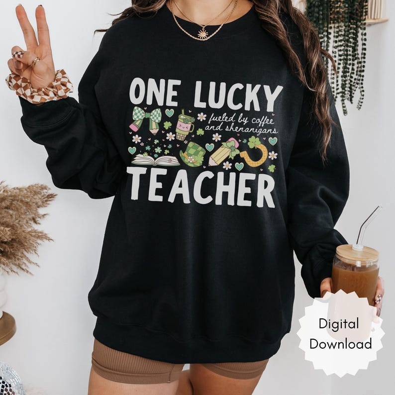 May include: Black sweatshirt with the text "ONE LUCKY TEACHER" in white, along with green and white graphics of a bow, coffee cup, shamrocks, and a pencil. The phrase "fueled by coffee and shenanigans" is also included.