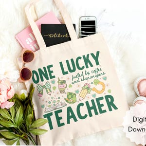 May include: Beige tote bag with the text "ONE LUCKY TEACHER" in green, along with St. Patrick's Day themed graphics. The bag is filled with a pink notebook, a black notebook, a phone, and sunglasses. A pair of pink sneakers is visible.