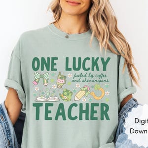 May include: Sage green t-shirt with a St. Patrick's Day theme. The shirt features the text "ONE LUCKY TEACHER" with graphics of a bow, coffee cup, pencil, and shamrock. The phrase "fueled by coffee and shenanigans" is also included.
