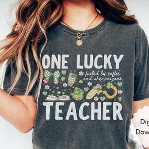 May include: Dark gray t-shirt with the text "ONE LUCKY TEACHER" and a graphic design featuring a bow, coffee cup, shamrocks, and a pencil. The shirt also includes the phrase "fueled by coffee and shenanigans."