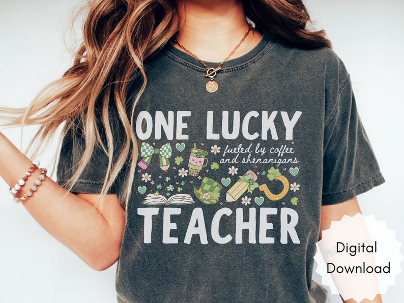 May include: Dark gray t-shirt with the text "ONE LUCKY TEACHER" and a graphic design featuring a bow, coffee cup, shamrocks, and a pencil. The shirt also includes the phrase "fueled by coffee and shenanigans."