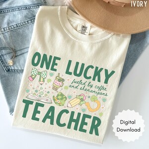 May include: Ivory-colored t-shirt with the text "ONE LUCKY TEACHER" in green, along with illustrations of a bow, pencil, coffee cup, and shamrock. The shirt also includes the phrase "fueled by coffee and shenanigans."