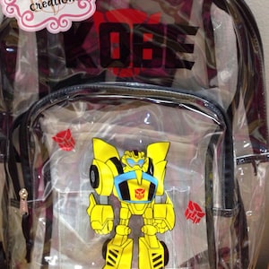 May include: A clear plastic backpack featuring a yellow and black robot character graphic on the front pocket. The name KOBE is printed in black letters above the robot. A red Autobot symbol is visible on the left side of the front pocket.