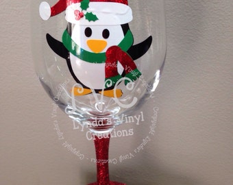 May include: A clear wine glass with a red glitter stem and base. The glass features a cartoon penguin wearing a Santa hat and scarf. The text "Lynda's Vinyl Creations" is printed on the glass.