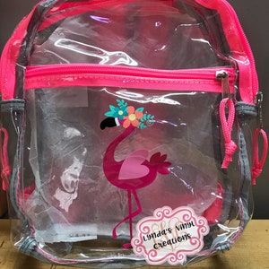 May include: Clear plastic backpack with a pink zipper and trim. A pink flamingo with a floral crown and a heart-shaped design is on the front of the bag. The bag also has a pink and white sticker with the text "Lynda's Vinyl Creations".