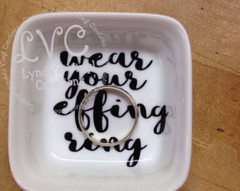 May include: White ceramic ring dish with black lettering that reads "wear your effing ring". A silver ring is sitting in the dish.