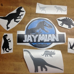 May include: A collection of dinosaur-themed decals in various shapes and sizes. Includes silhouettes of a T-Rex, Stegosaurus, and Brachiosaurus, along with a logo that reads "JAYMIAN". The decals are in shades of black and gray.