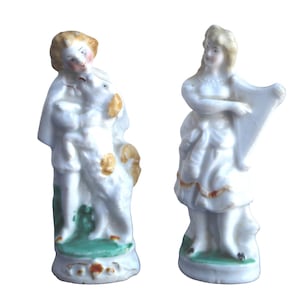 May include: Two antique porcelain figurines. One figure is a man in white clothing with gold accents, holding a sheep. The other is a woman in a white dress playing a harp. Both figures have a green base.