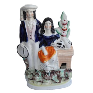 May include: A vintage ceramic figurine depicting a couple in period clothing. The man wears a blue jacket and white breeches, holding a hat. The woman is in a blue dress, holding an orange fan. A dog rests near a small structure.