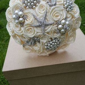 May include: A white satin bridal bouquet decorated with pearl and rhinestone embellishments, including a starfish, a seahorse, and other decorative elements.