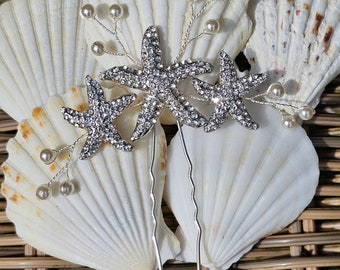 May include: Two silver hair pins with rhinestone starfish and pearl accents. The pins are displayed on a seashell background.