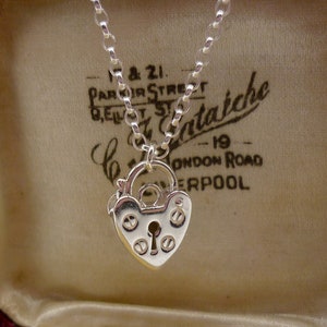 Può includere: Una collana d'argento con un ciondolo a forma di lucchetto a forma di cuore, con un buco della serratura e dettagli decorativi. La collana è esposta in una scatola per gioielli in velluto rosso con fodera in raso bianco. La scatola ha un'etichetta che recita "1 & 21, Parker Street, Belfast, Co.Antrim, London Road, Liverpool."