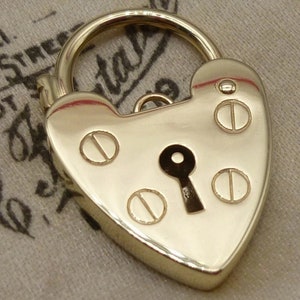 May include: A gold heart-shaped padlock with a keyhole and decorative accents. The padlock is on a white fabric surface.