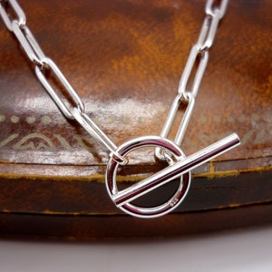 May include: A silver necklace with a toggle clasp. The chain is made of elongated links, and the clasp features a circular ring and a straight bar. The jewelry is displayed on a brown surface.